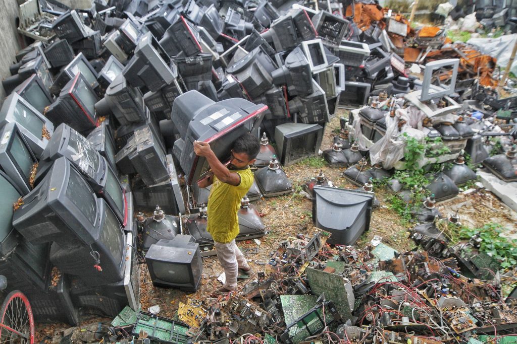 Man carrying an old television through a scrapyard filled with discarded electronics and piles of broken devices.